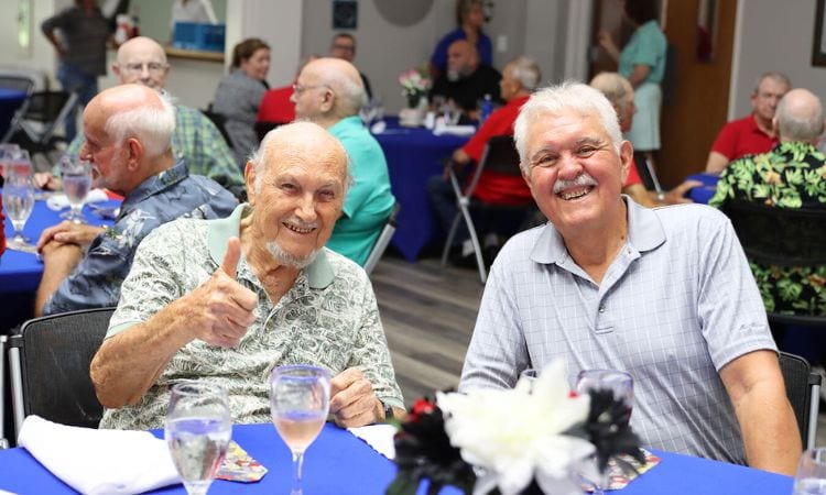 Two senior men are seated at a table. Both smile. One gives a 'thumbs up.'