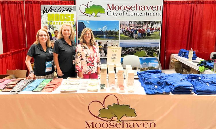 Three women stand behind table in front of Moosehaven banner