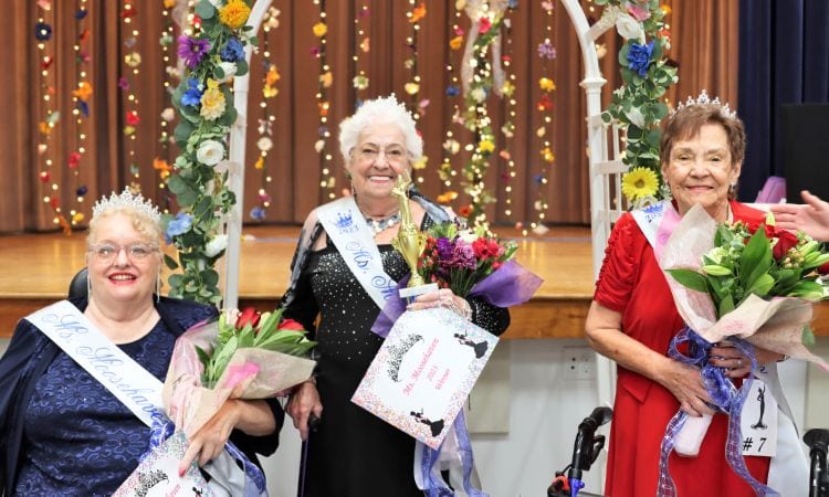 Three senior ladies in fancy gowns, tiaras, and sashes hold flowers